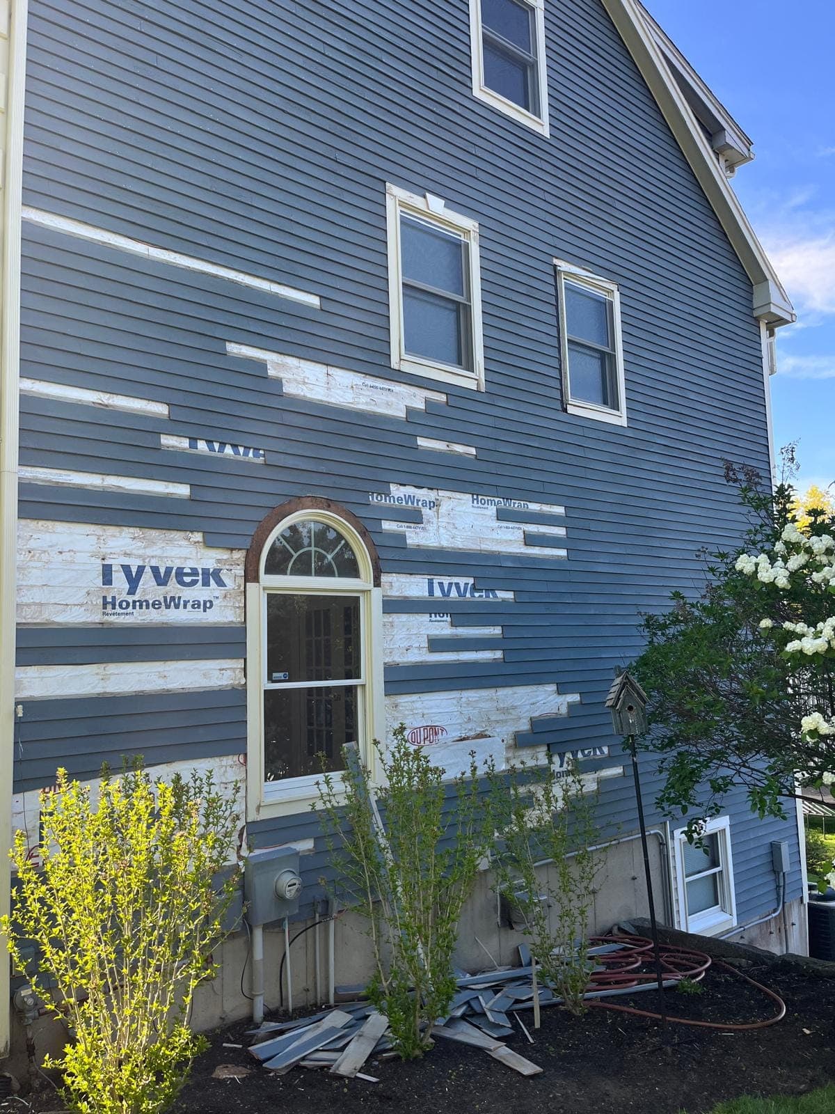 Front of a blue clapboard house with runs of siding removed and house wrap showing around a round-top window
