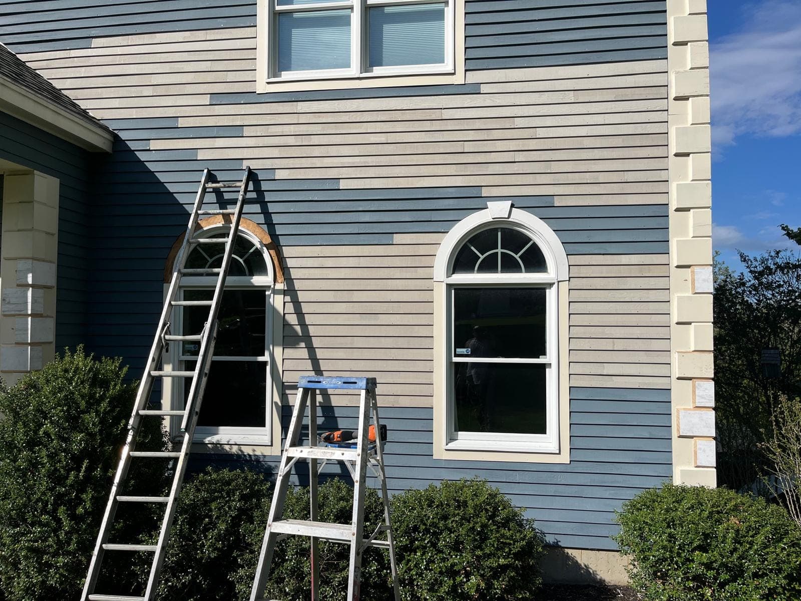 Two ladders against the gable end of a blue clapboard house, a round-top window above